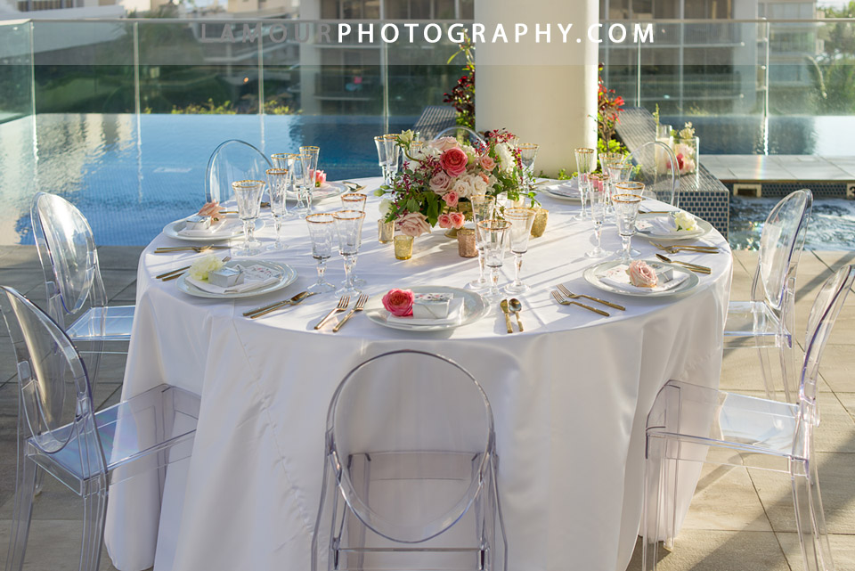 Oahu wedding reception at a hotel in Honolulu with clean white table cloth and clear oval back chairs with large floral centerpiece and gold decor accents