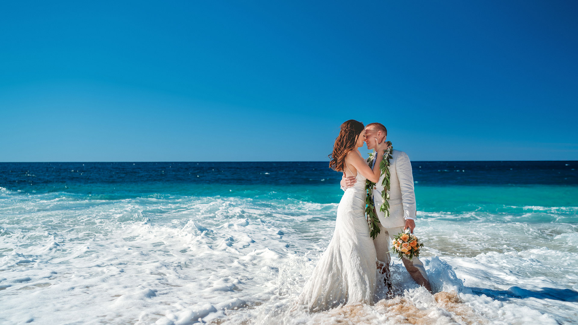 A wave washes around a wedding couple as they kiss