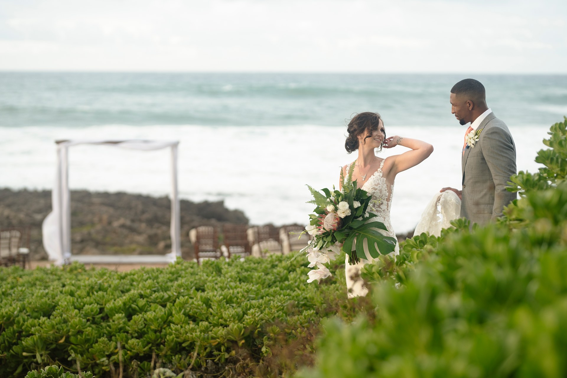 Bride and groom sharing a look among coastal greenery, with an oceanfront ceremony site behind them
